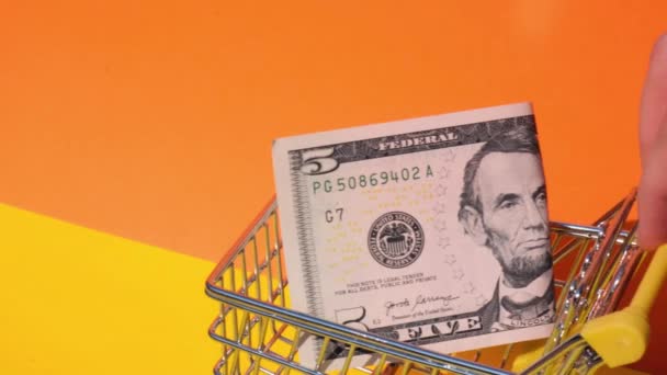 Hand pushing toy supermarket trolley with 5 dollar banknote Money in shopping cart on yellow background. Sales Basket of US dollar bills. Minimum living wage Concept: loan, investment pension saving - Footage, Video