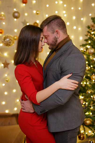 Nettes Paar in festlicher Kleidung umarmt in weihnachtlich dekoriertem Zimmer - Foto, Bild
