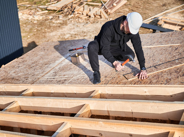 Carpenter hammering nail into OSB panel on the roof top of future cottage. Man worker building wooden frame house. Carpentry and construction concept. - Foto, afbeelding