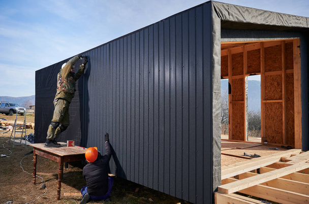 Male builders installing black corrugated iron sheet used as facade of future cottage. Men workers building wooden frame house in the Scandinavian style barnhouse. Carpentry and construction concept. - Foto, Imagem