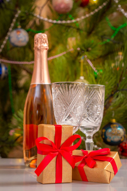 ギフトボックスとぼやけたクリスタルグラス、シャンパンのボトル。背景にパーティーライトやおもちゃのボールがちらつきクリスマスツリー. - 写真・画像