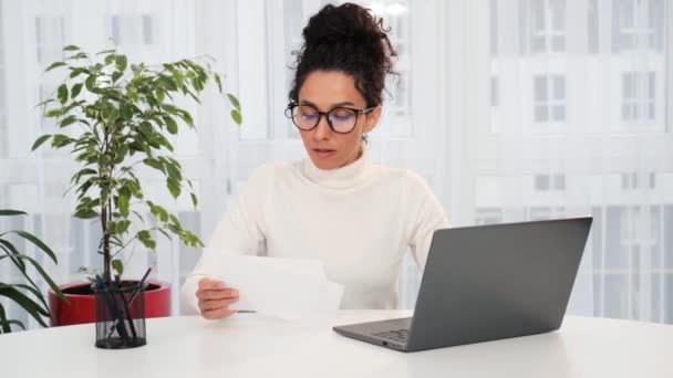 Latynoska kobieta w okularach siedząca na stole, zarabiająca rachunki za pomocą laptopa, kontrolująca budżet, kontrolująca zaległe spłaty pożyczek, czuje się zestresowana w domu. Kryzys, brak pieniędzy, koncepcja zadłużenia - Materiał filmowy, wideo