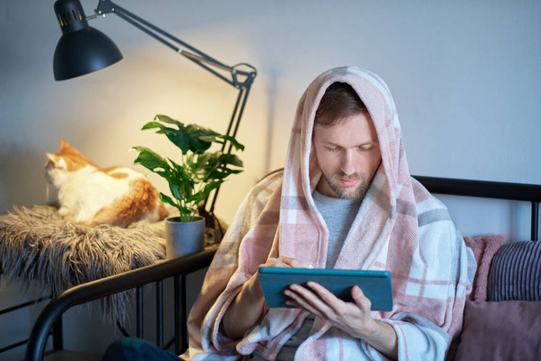 Hombre caucásico freelancer cubierto con manta usando tableta digital en la cama y gato tabby blanco sentado cerca de él. Trabajo a distancia de la oficina en casa o la educación en línea. Sensación de malestar o enfermedad - Foto, Imagen