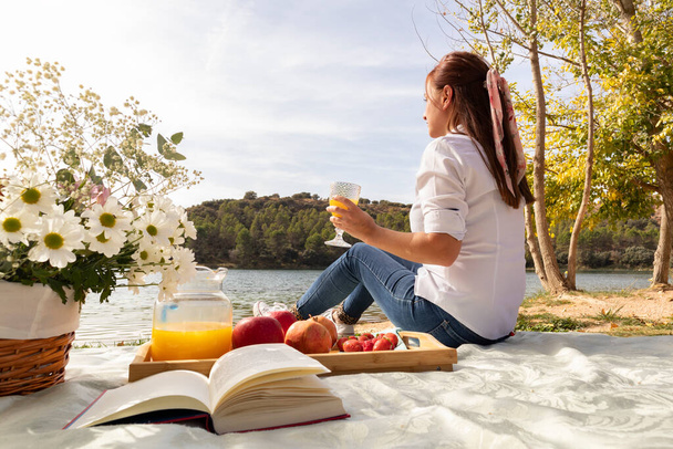 дівчина читає книгу на пікніку з квітами та фруктами на березі озера
 - Фото, зображення