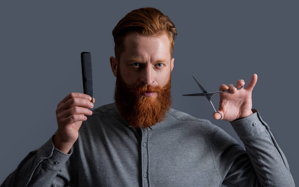 man holding barber scissors and comb. barber with hairstyle. barber man making hairstyle isolated on grey background. barber man with scissors do hairstyle. man has beard and hairstyle in studio. - Photo, Image