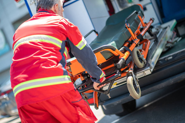 Back view of a gray-haired paramedical worker pulling the gurney out of the ambulance van - Fotoğraf, Görsel