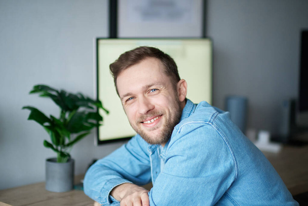 Cute attractive caucasian man in denim shirt with positive emotion and computer on background. Cheerful bearded male freelancer sitting in home office looking at camera smiling. High quality image - Foto, Imagem