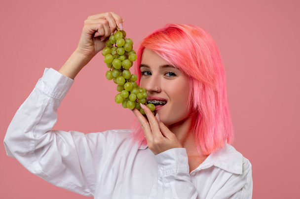 Portrait of a girl with the metal braces biting a berry off a bunch of grapes in her hands - Photo, Image