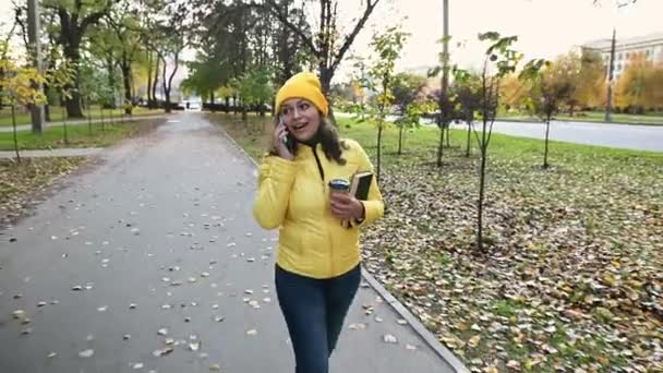 Jolie femme bouclée aux cheveux bruns et multi-ethnique avec un chapeau et une veste jaune vif, parlant sur un téléphone portable tout en marchant dans la rue avec des feuilles d'automne, par une chaude journée d'automne ensoleillée. L'été indien. Novembre - Séquence, vidéo