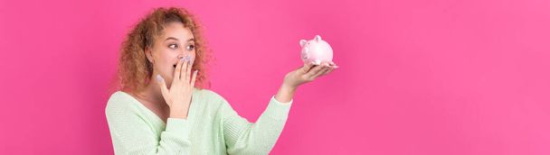 A cute young girl with curly red hair holds a piggy bank, a pink piglet in her hands. The concept of wealth and safety of money. - Zdjęcie, obraz