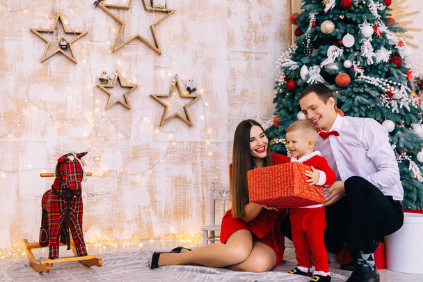 bébé en costume de Père Noël tient grand coffret cadeau et sourire. parents assis près de l'arbre de Noël et souriant. cheval à bascule dans la chambre. - Photo, image