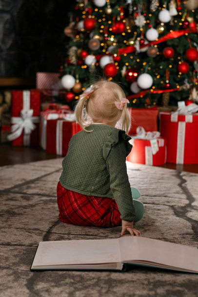 Kleines Mädchen sitzt auf dem Boden der Reihe mit großem Buch und blickt auf schönen Weihnachtsbaum. Rückansicht. Vertikaler Rahmen - Foto, Bild