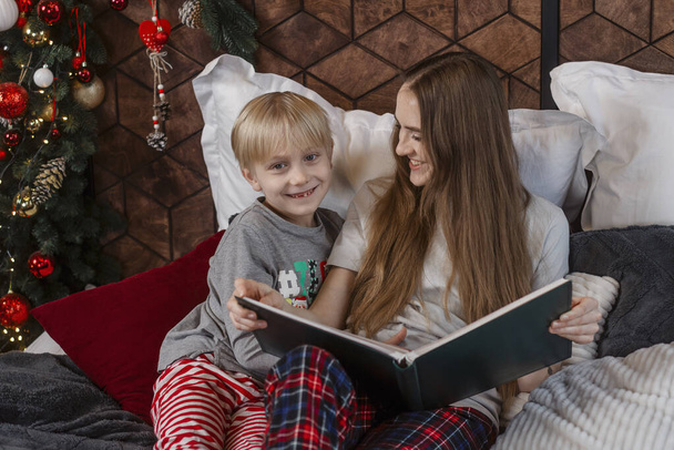 Mother telling funny fairy tale to cute blond boy. Mother and child son reading book lying in bed near Christmas tree - Фото, зображення