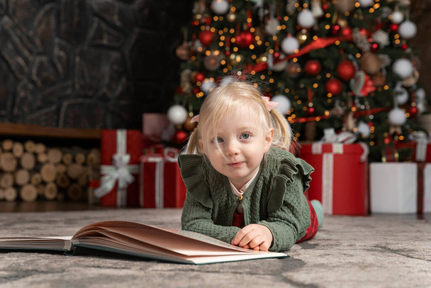 Ritratto di graziosa bambina bionda sdraiata sul pavimento accanto a un grande libro sullo sfondo dell'albero di Natale. Bambini e umore di Capodanno - Foto, immagini