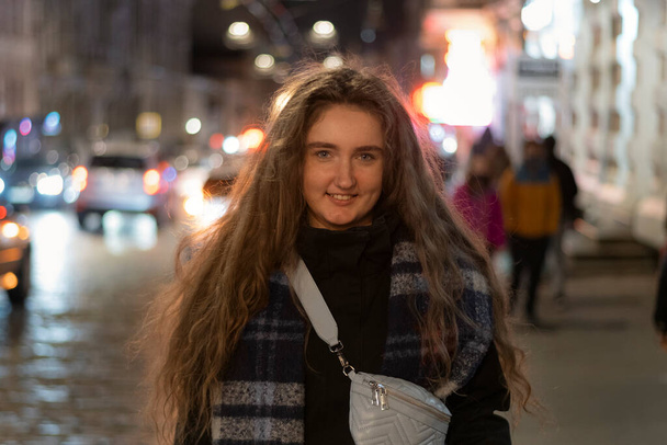 Молодая улыбающаяся девушка с длинными пышными каштановыми волосами в осеннем пальто на улице ночного города - Фото, изображение