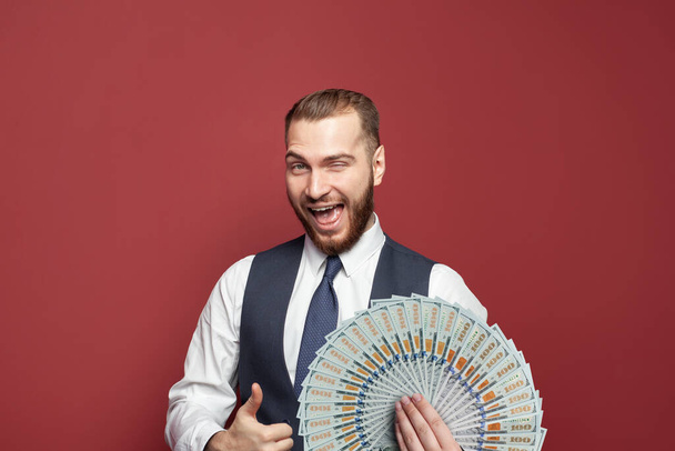 Excited successful businessman winking and showing thumb up with money us dollars fan on red wall background - Photo, Image