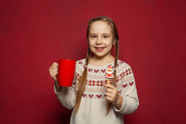 Christmas portrait of happy child girl with red mug and Santa Claus cookie on red background - Photo, Image