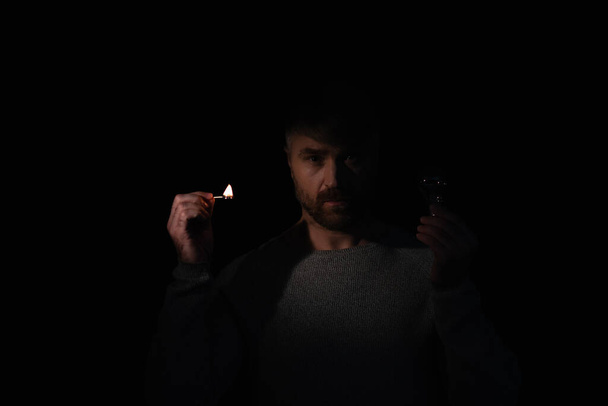 Mann mit Glühbirne und brennendem Streichholz blickt isoliert auf schwarze Kamera - Foto, Bild