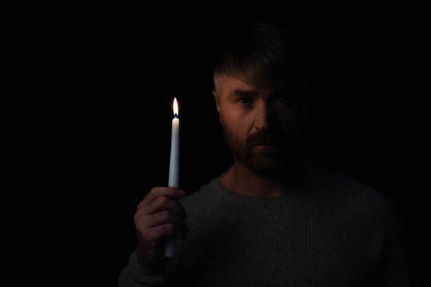 hombre sosteniendo vela encendida durante el apagón de energía y mirando a la cámara aislada en negro - Foto, Imagen