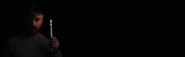 άνθρωπος κρατώντας αναμμένο κερί και κοιτάζοντας κάμερα κατά τη διάρκεια της διακοπής ρεύματος απομονώνονται σε μαύρο, πανό - Φωτογραφία, εικόνα