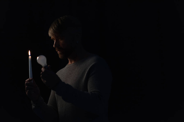 man holding light bulb and burning candle in darkness isolated on black - Photo, Image