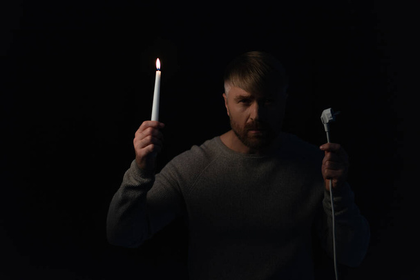 man with plug and burning candle looking at camera during electricity shutdown isolated on black - Photo, Image