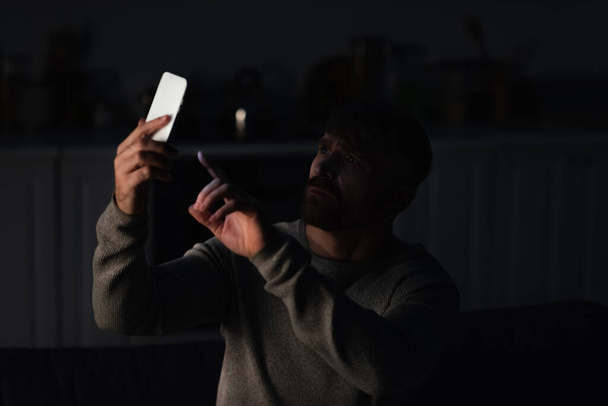 homme inquiet pointant vers le téléphone portable tout en cherchant une connexion mobile pendant une panne d'électricité - Photo, image