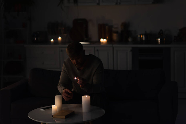 άνθρωπος κάθεται σε σκοτεινή κουζίνα κοντά στην καύση κεριών και βιβλίο με smartphone  - Φωτογραφία, εικόνα