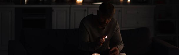 muž se zapálenou zápalkou sedí v kuchyni poblíž svíček při výpadku energie, banner - Fotografie, Obrázek