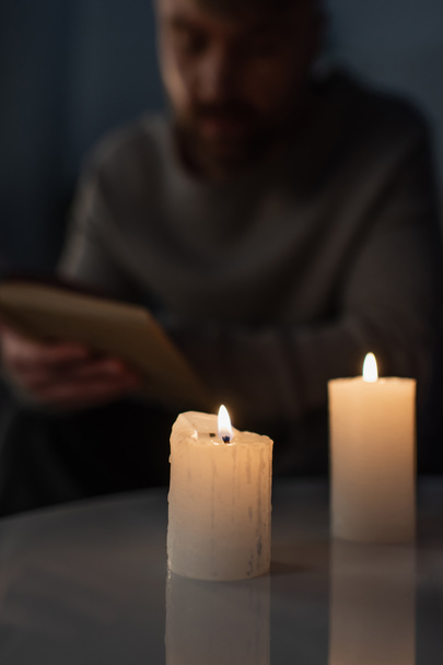wazig man lezen boek in duisternis in de buurt van brandende kaarsen tijdens energie black-out - Foto, afbeelding