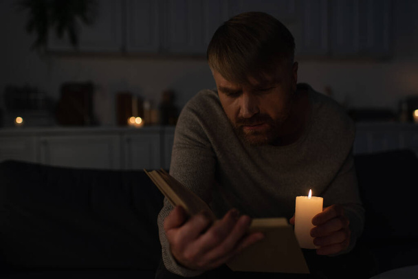 man holding lit candle while reading book in dark kitchen during electricity outage - Photo, Image