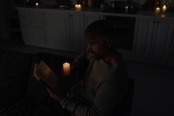 high angle view of man reading book during electricity shutdown in kitchen with burning candles  - Foto, Bild