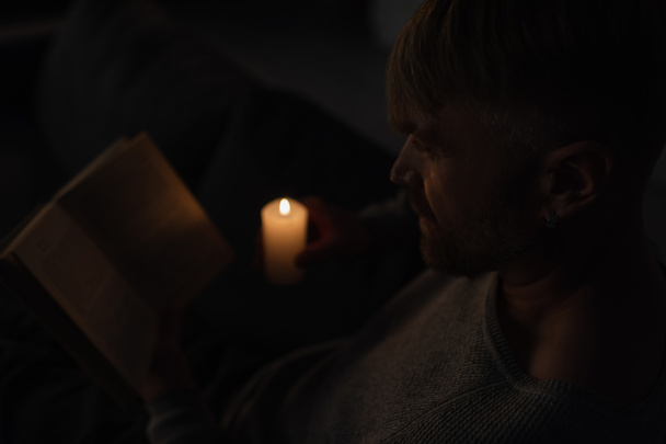 hombre sosteniendo vela encendida mientras lee libro en la oscuridad causada por apagón de energía - Foto, Imagen