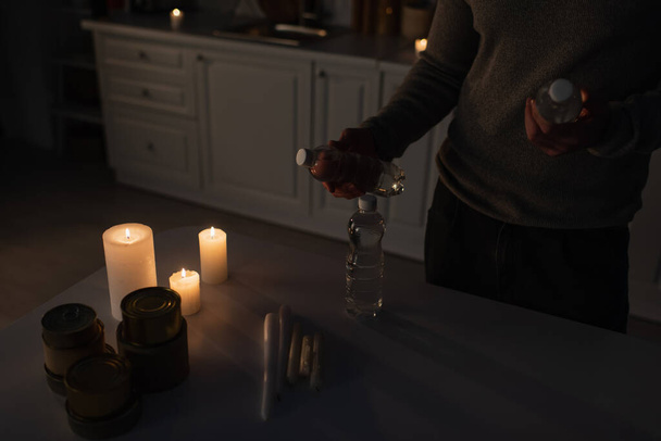 cropped view of man with bottled water near table with canned food and candles in dark kitchen - Foto, imagen