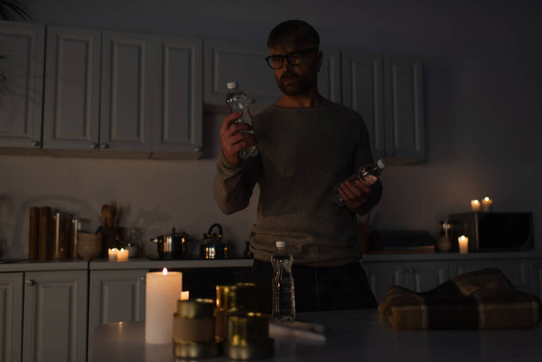 mężczyzna w okularach trzyma butelkowaną wodę przy stole z konserwami i ciepłym kocem w ciemnej kuchni - Zdjęcie, obraz