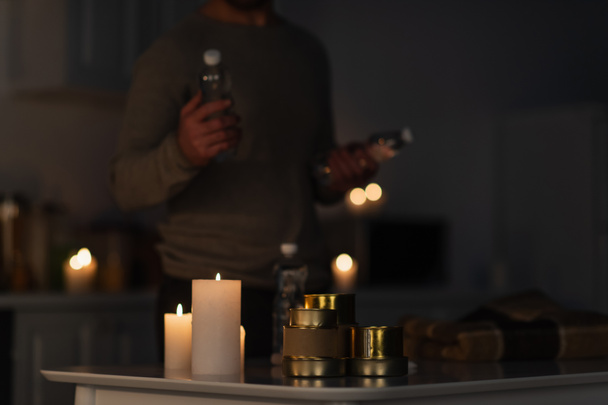 cropped view of blurred man holding bottled water near table with stock of canned food near candles and warm blanket - Fotoğraf, Görsel