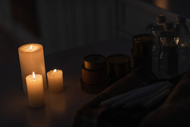 консервы и бутилированная вода возле теплого одеяла и зажженных свечей во время отключения энергии - Фото, изображение