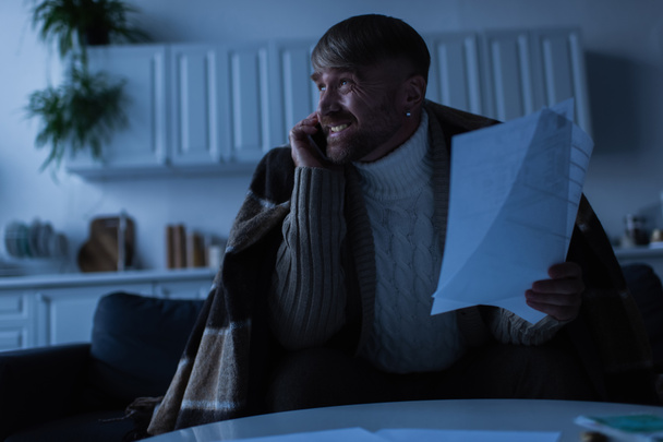 nervoso e congelado homem falando no smartphone enquanto segurando contas de pagamento no crepúsculo  - Foto, Imagem