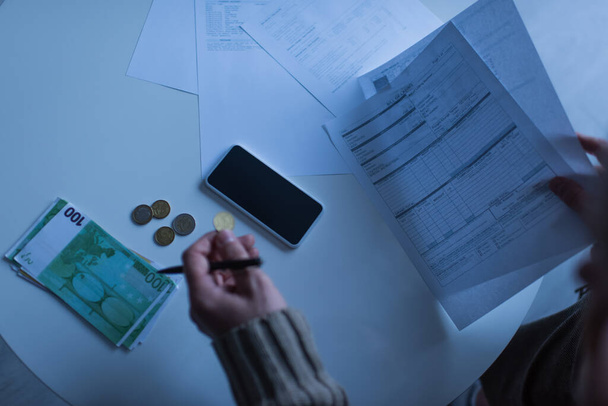 KYIV, UKRAINE - NOVEMBER 2, 2022: partial view of man holding invoices near money and mobile phone in twilight - Φωτογραφία, εικόνα