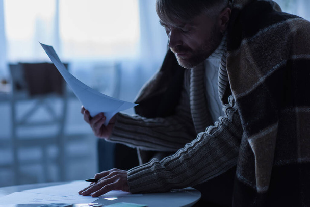congelado hombre cubierto con manta caliente mirando facturas de pago en crepúsculo - Foto, Imagen