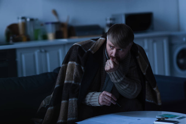 frozen man looking away while sitting under blanket near invoices and money during power blackout - Foto, imagen