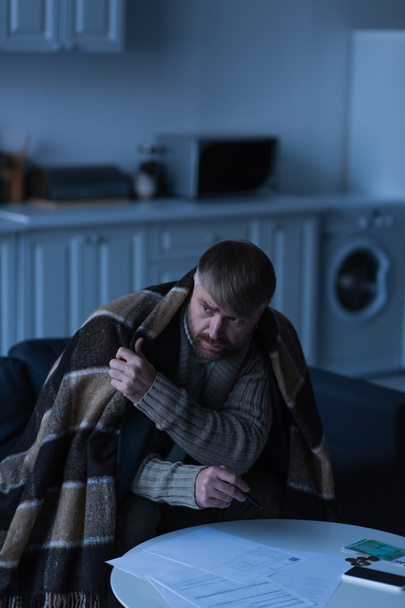 upset and frozen man sitting under warm blanket near invoices and money in twilight - Foto, imagen