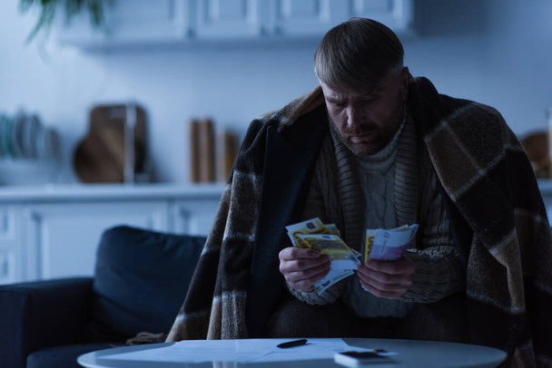homme assis sous couverture et comptant de l'argent près des factures pendant la panne d'énergie - Photo, image