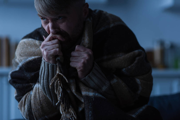 homem estressado segurando a mão perto do rosto enquanto sentado sob cobertor quente e olhando para longe no crepúsculo - Foto, Imagem