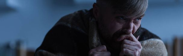 homem deprimido sentado sob cobertor quente e segurando a mão perto do rosto durante o desligamento da eletricidade, banner - Foto, Imagem