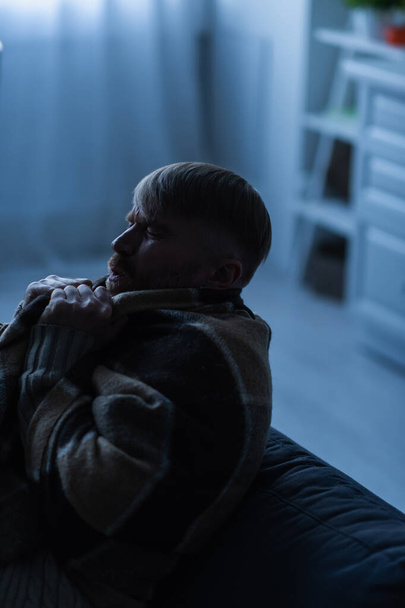 hombre congelado con los ojos cerrados sentado en casa bajo una manta caliente durante el apagón eléctrico - Foto, imagen