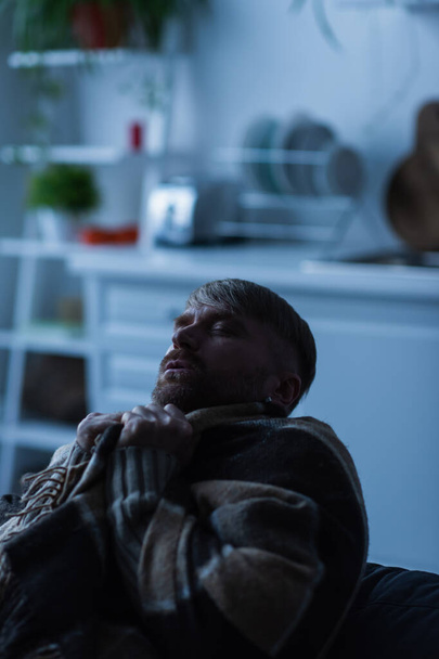 Sıcak battaniyeye sarılı donmuş bir adam bulanık bir mutfakta gözleri kapalı oturuyor.  - Fotoğraf, Görsel