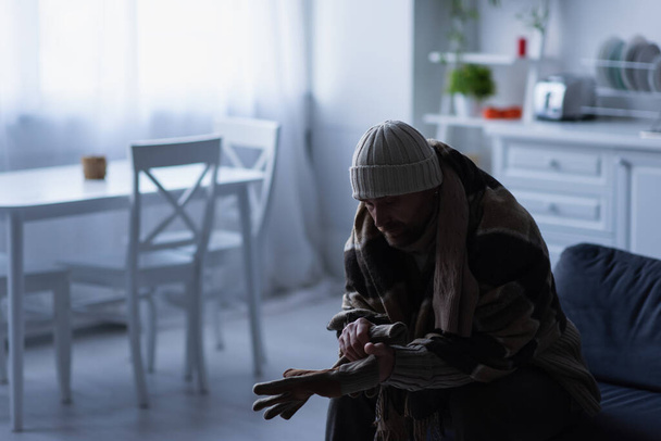 κατεψυγμένος άνδρας με πλεκτό καπέλο κάθεται στην κουζίνα κάτω από κουβέρτα και φοράει ζεστά γάντια - Φωτογραφία, εικόνα