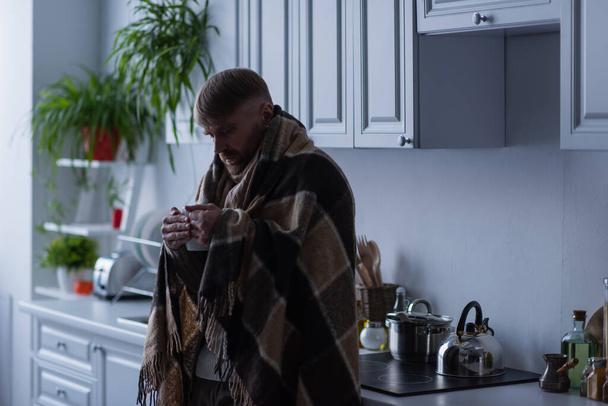Donmuş adam mutfakta battaniyenin altında duruyor ve elinde bir fincan sıcak çay tutuyor. - Fotoğraf, Görsel