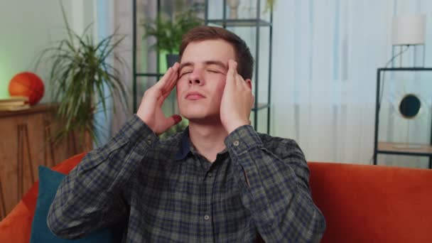 Hombre adolescente caucásico disgustado frotando las sienes para curar el problema del dolor de cabeza, que sufre de tensión y migraña, estrés, muecas de dolor, presión arterial alta. Chico joven en casa sala de estar en el sofá - Imágenes, Vídeo
